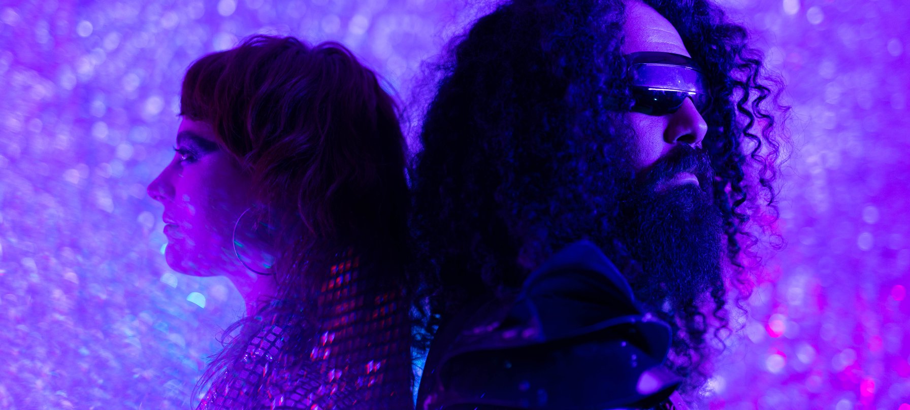 A photo of two Golden Sunbird members washed with sparkly purple lights making it look like they are on a dance floor. The members stand back-to-back wear reflective clothing. Credit Gabrielle Eve.