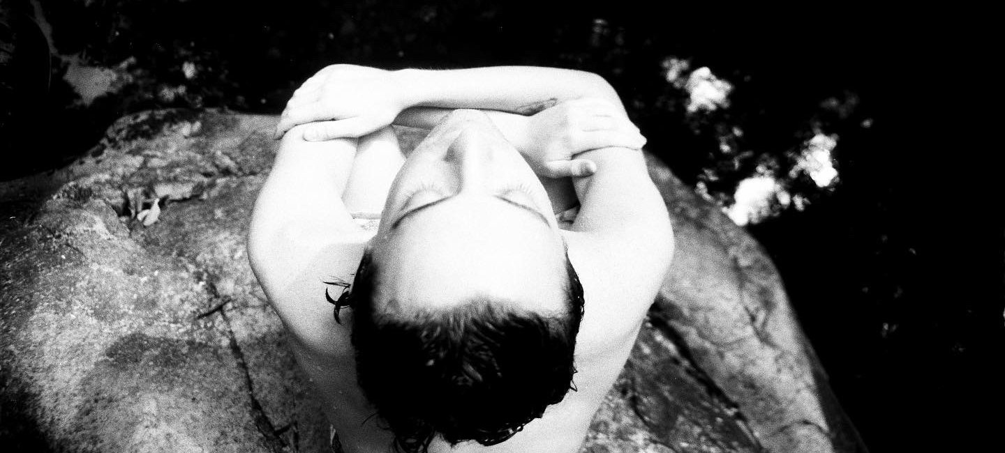 A black and white photo taken from a bird's eye view of Pierce Eldridge. Pierce holds their knees to their chest atop a rock surrounded by water. Credit courtesy of artist.