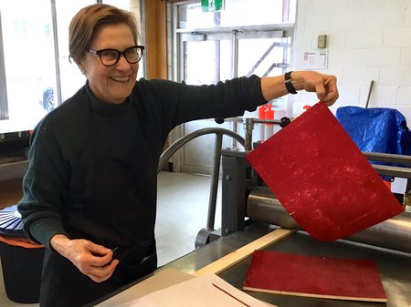 A photo of Helen Mueller with a big smile facing the camera. Helen holds up a sheet of paper that has just gone through the printing press. The sheet is covered in red ink with gentle carvings of white paper showing throughout. Credit Helena Demzcuk.