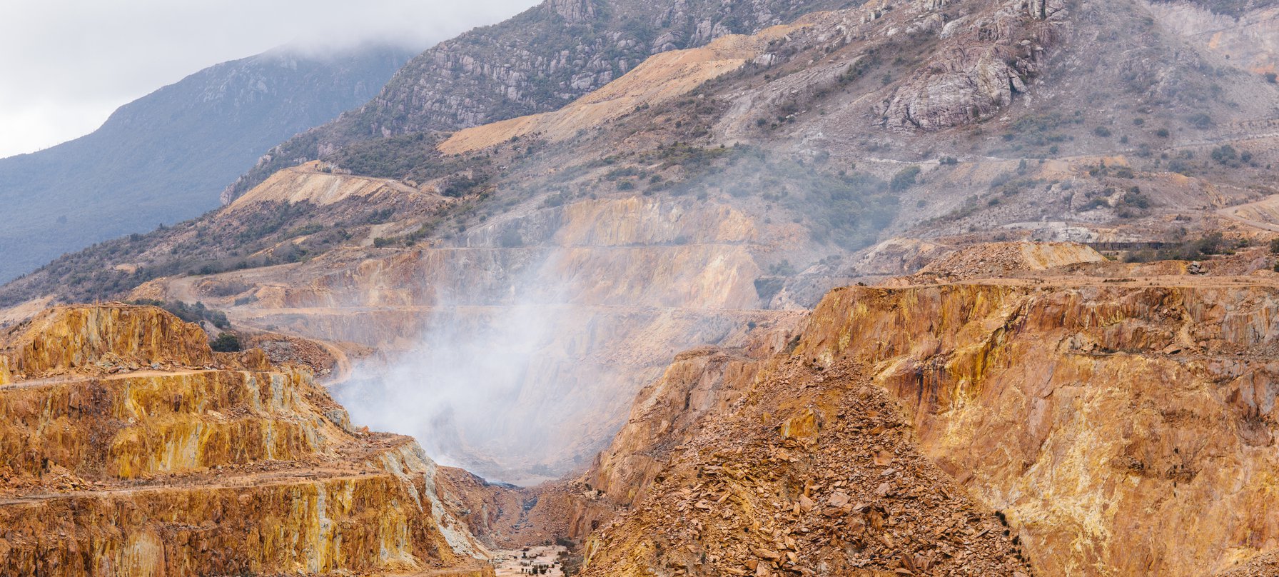 A photo of smoke rising from a terraced mine site at the Mt Lyell mine, with rocky Mt Lyell in the background. Image by Jesse Hunniford.