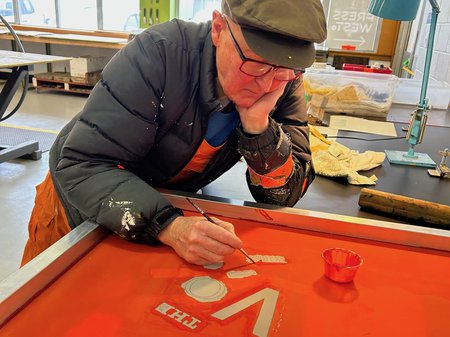 A photo of Raymond Arnold at work. Raymond is leaning on a workbench with his head resting on his hand. Raymond is painting lettering with a rich orange colour. Raymond has pale skin, wide rectangle glasses and is wearing a flat cap. Image Raymond Arnold.