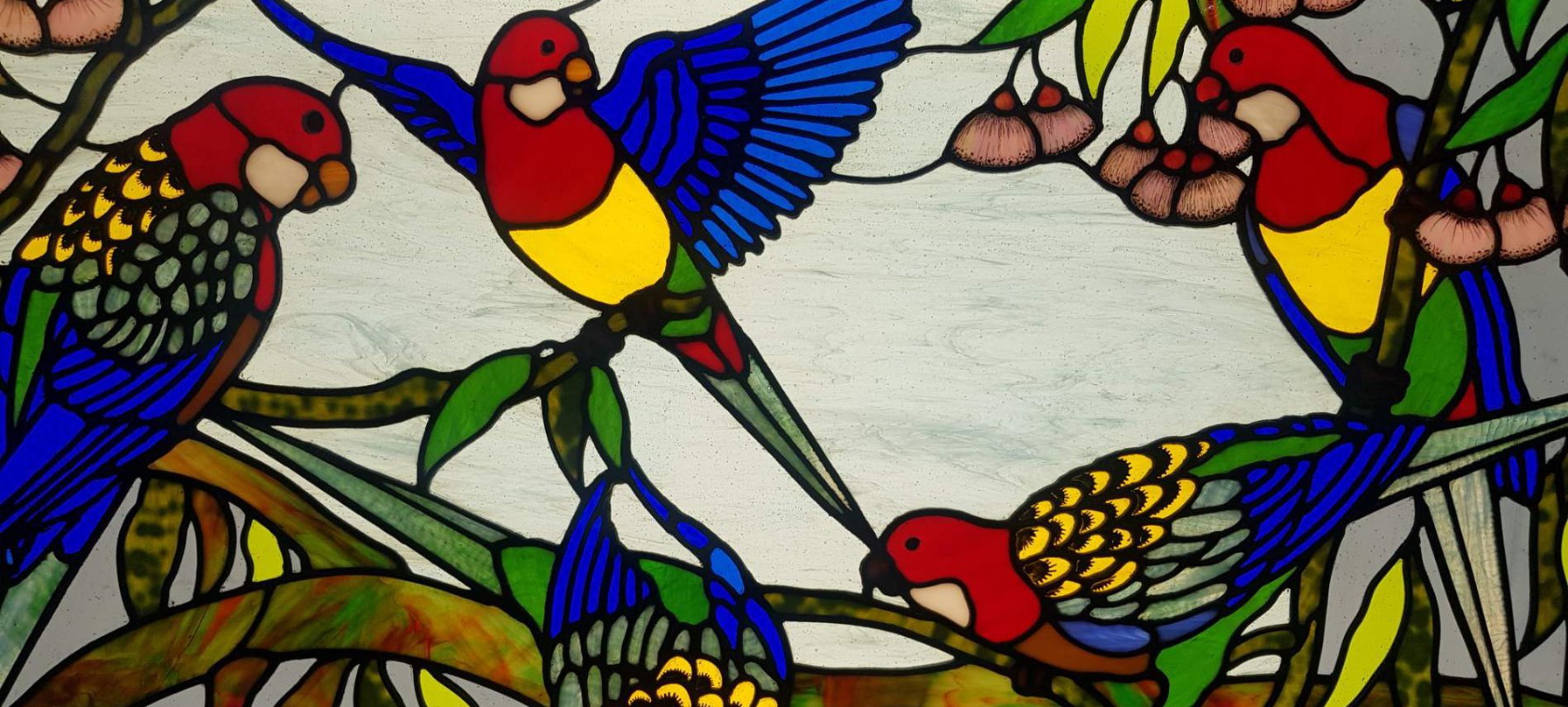 A photo of Brenda Steiner's stained glass art. The work features a family of Rosella birds. The birds are gathered on a tree branch, some flapping their wings and others pecking at food. Credit Brenda Steiner.