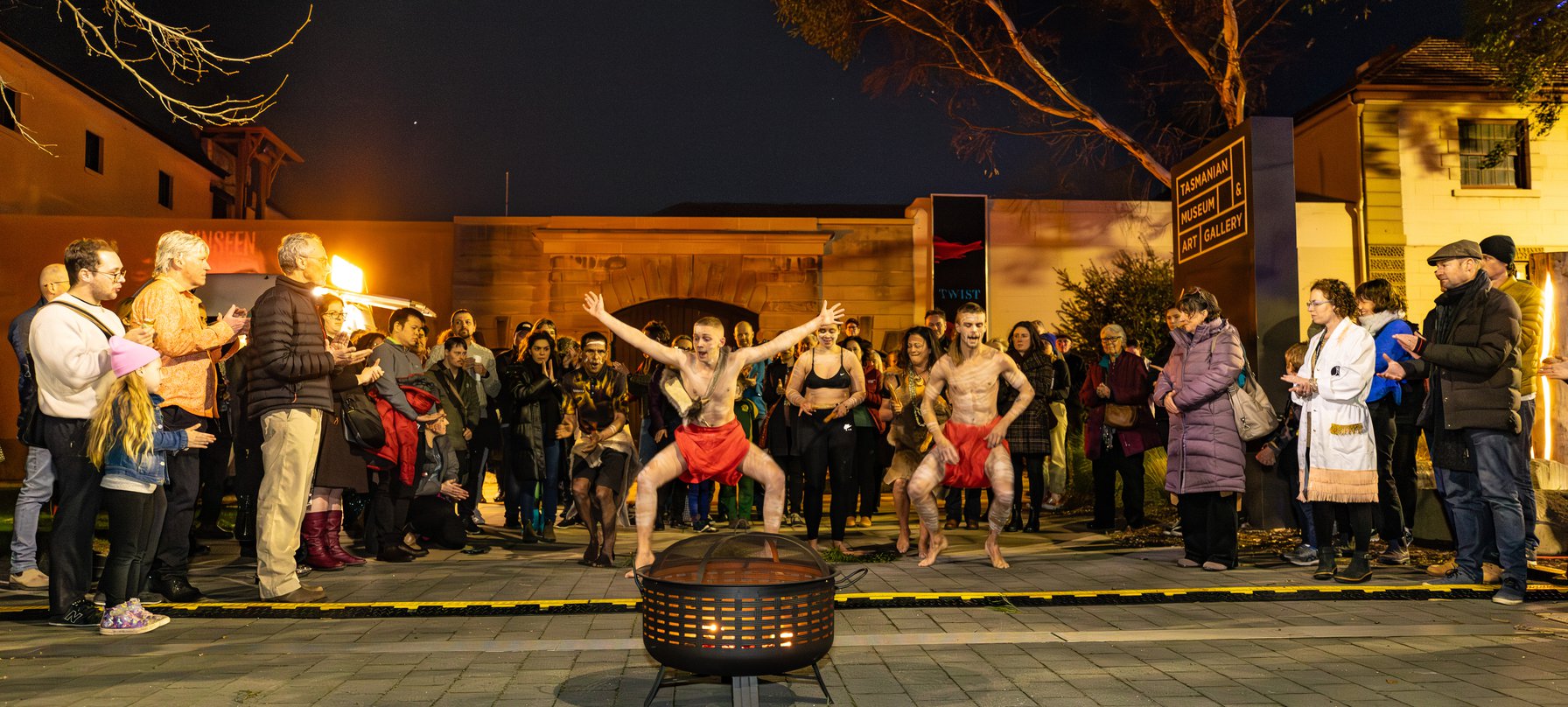 A photo of Sienna, Zane and Xavier Scotney-Barron performing traditional Aboriginal dance and song around a fire pit. Performers are leaping in the air while other use clap sticks. A crowd encircles them, watching. Credit Jillian Mundy.