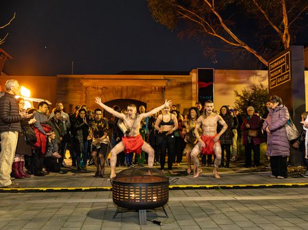 A photo of Sienna, Zane and Xavier Scotney-Barron performing traditional Aboriginal dance and song around a fire pit. Performers are leaping in the air while other use clap sticks. A crowd encircles them, watching. Credit Jillian Mundy.