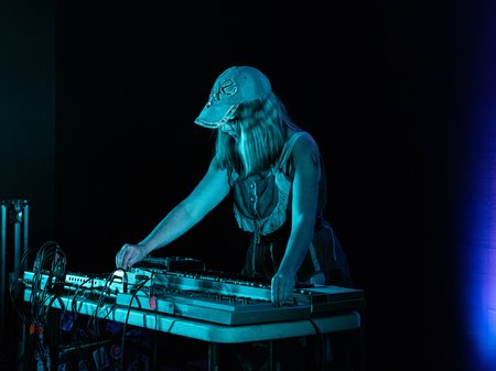 Artist SCRAPS stands behind a DJ deck in a dark room, looking down at the controller as blue lighting illuminates them.