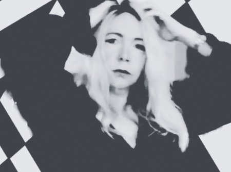 Artist Scratch Kelly stands in front of a black-and-white checkered wall, looking directly into the camera with their hands in their hair.
