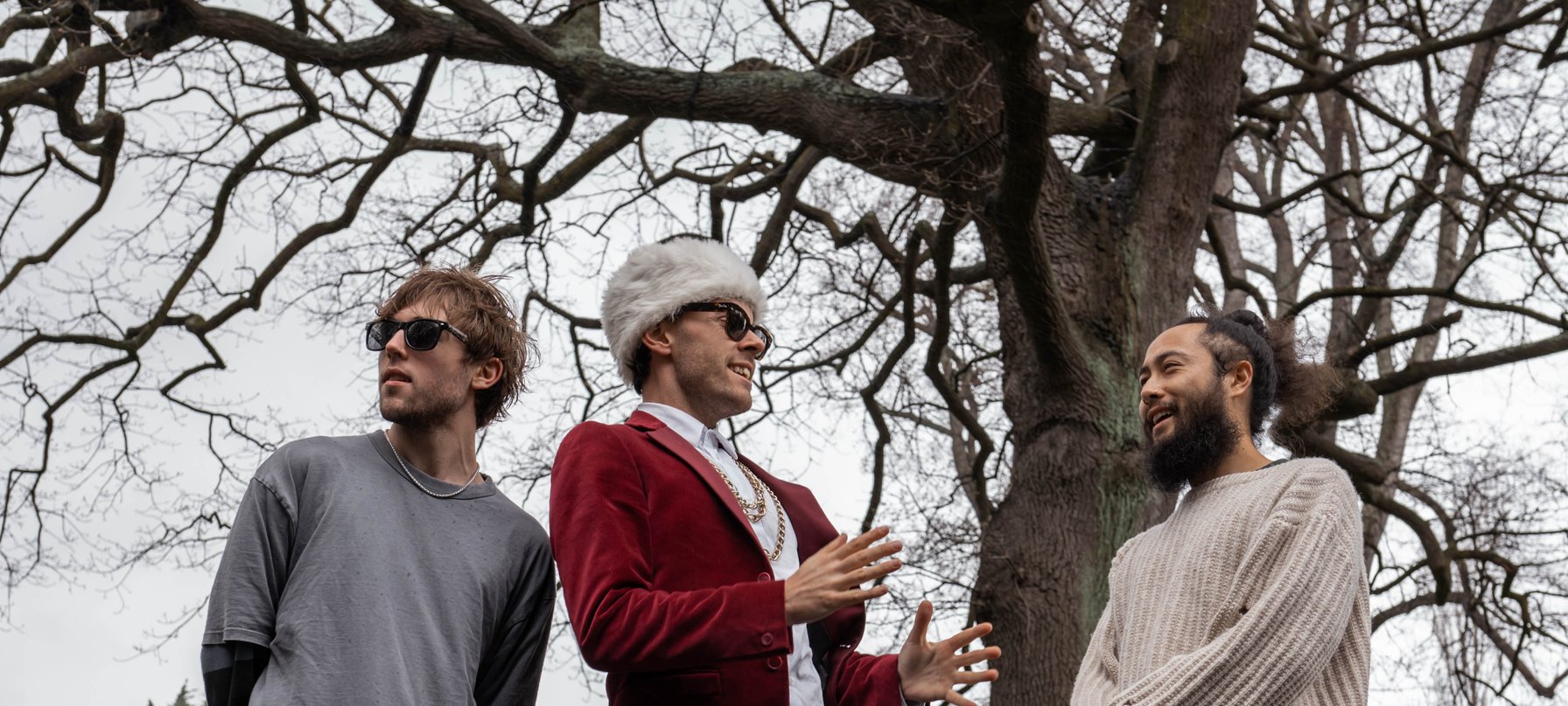 A photo of the 3 Slaughterhäus Surf Cult band members. 2 people wear sunglasses. 1 wears a fluffy hat and a gold chain. The background is a tall tree with no leaves. Credit Mell Schmeider.