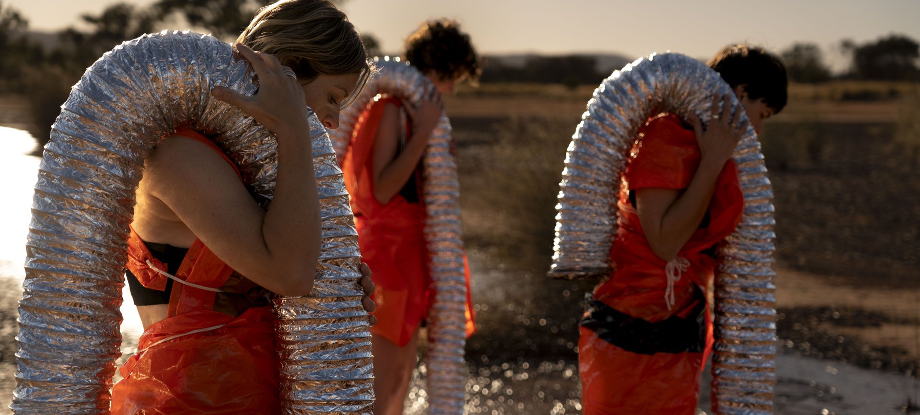 SUB image. A photo of three people dressed in orange plastic with lengths of flexible foil duct carried over their shoulders. These people are walking through a wet, gravelly terrain. Credit Ivan Trigo Miras.