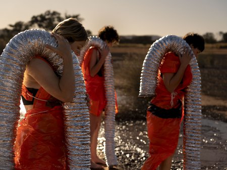 SUB image. A photo of three people dressed in orange plastic with lengths of flexible foil duct carried over their shoulders. These people are walking through a wet, gravelly terrain. Credit Ivan Trigo Miras.