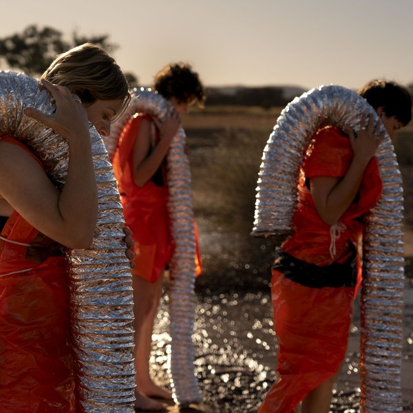 SUB image. A photo of three people dressed in orange plastic with lengths of flexible foil duct carried over their shoulders. These people are walking through a wet, gravelly terrain. Credit Ivan Trigo Miras.