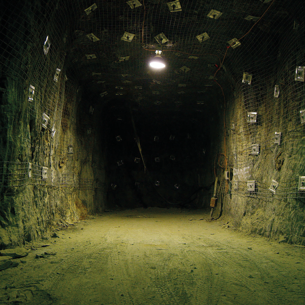 Thresholds. A photo in the Mount Lyell underground. Facing into a dim-lit tunnel, the photo captures raw rock walls covered in caging and a dirt floor with overlapping tyre marks. Credit Polly Stanton.