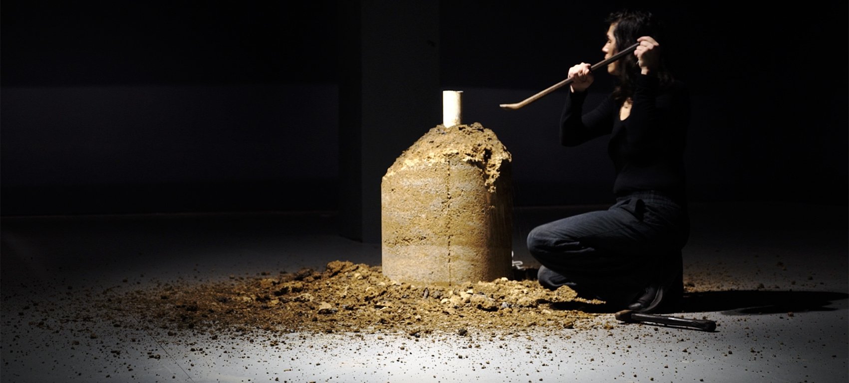 A photo of Selena de Carvalho deinstalling her past work named 'Readymade Burnouts. Selena is kneeling on the ground in a darkened room and using a crowbar to break down a rammed earth pillar. Credit courtesy of artist.