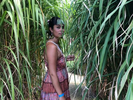 Tasha Jean stands among long, glossy green leaves, wearing a pink patterned dress and partially hidden by the foliage as they look over their shoulder.