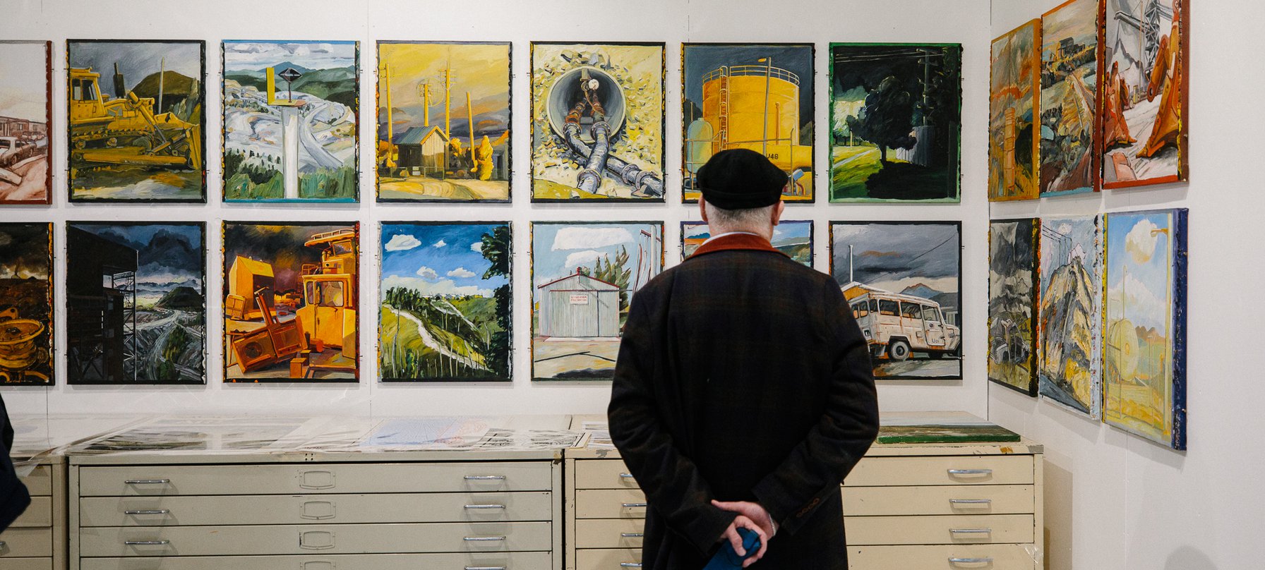 A photo of a man, dressed in black with his hands clasped behind his back, looking at artwork on a wall. The artwork is vibrant painted depictions of industrial landscape scenes. Image by Jesse Hunniford.