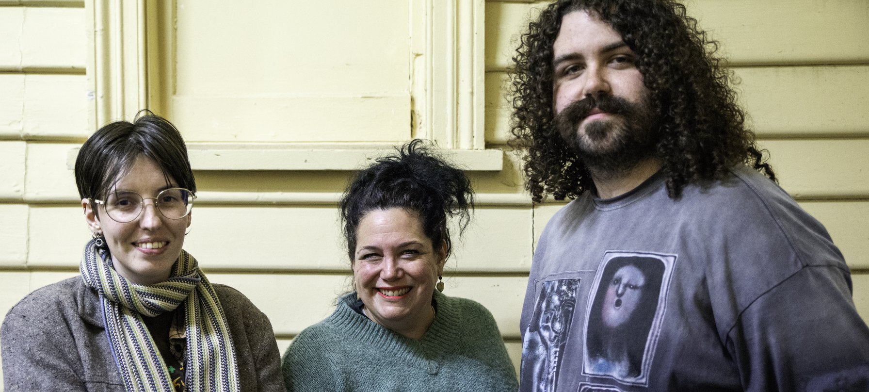 A photo of three people - The Unconformity's Ongoing Artists, Iris Blazely, Nancy Mauro-Flude and Stevie Battese - standing in a row against a yellow wooden building.