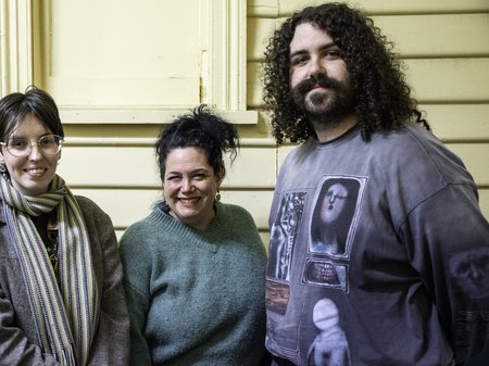A photo of three people - The Unconformity's Ongoing Artists, Iris Blazely, Nancy Mauro-Flude and Stevie Battese - standing in a row against a yellow wooden building.