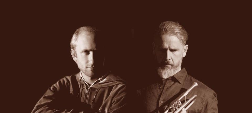 A photo of the two artists of GROUND. The two people are shadowed by a deep dark background. One holds a trumpet, the other leans of a steel guitar.