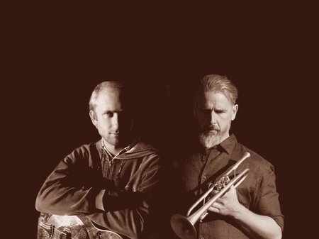 A photo of the two artists of GROUND. The two people are shadowed by a deep dark background. One holds a trumpet, the other leans of a steel guitar.