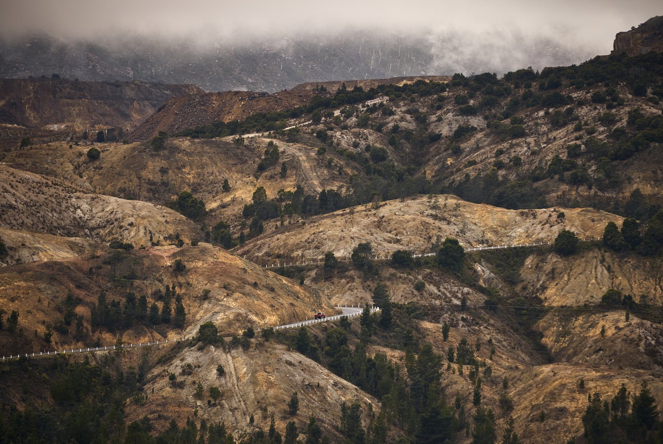 A photo of the hills surrounding Queenstown, lutruwita/Tasmania. Rocky orange hills are dotted with trees, with a grey road winding through the hills. Image by Rémi Chauvin.