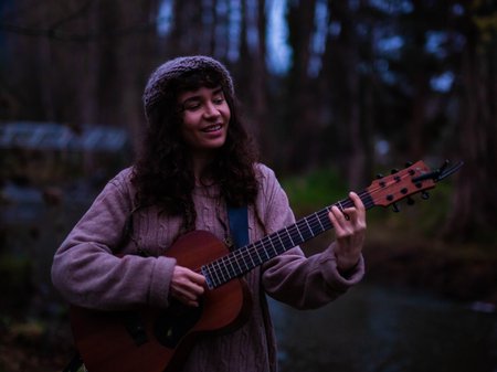A photo of Tasha Zappala smiling as she plays the guitar in a a wet forest area. Tash has long curly brown hair and tanned skin. She wears a woollen beanie, woollen jumper and long skirt. Credit courtesy of artist.