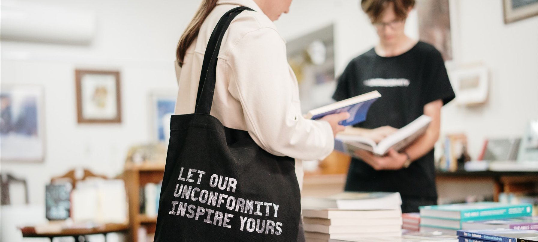Image of person carrying a The Unconformity tote bag and reading a book in a bookshop.