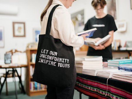 Image of person carrying a The Unconformity tote bag and reading a book in a bookshop.