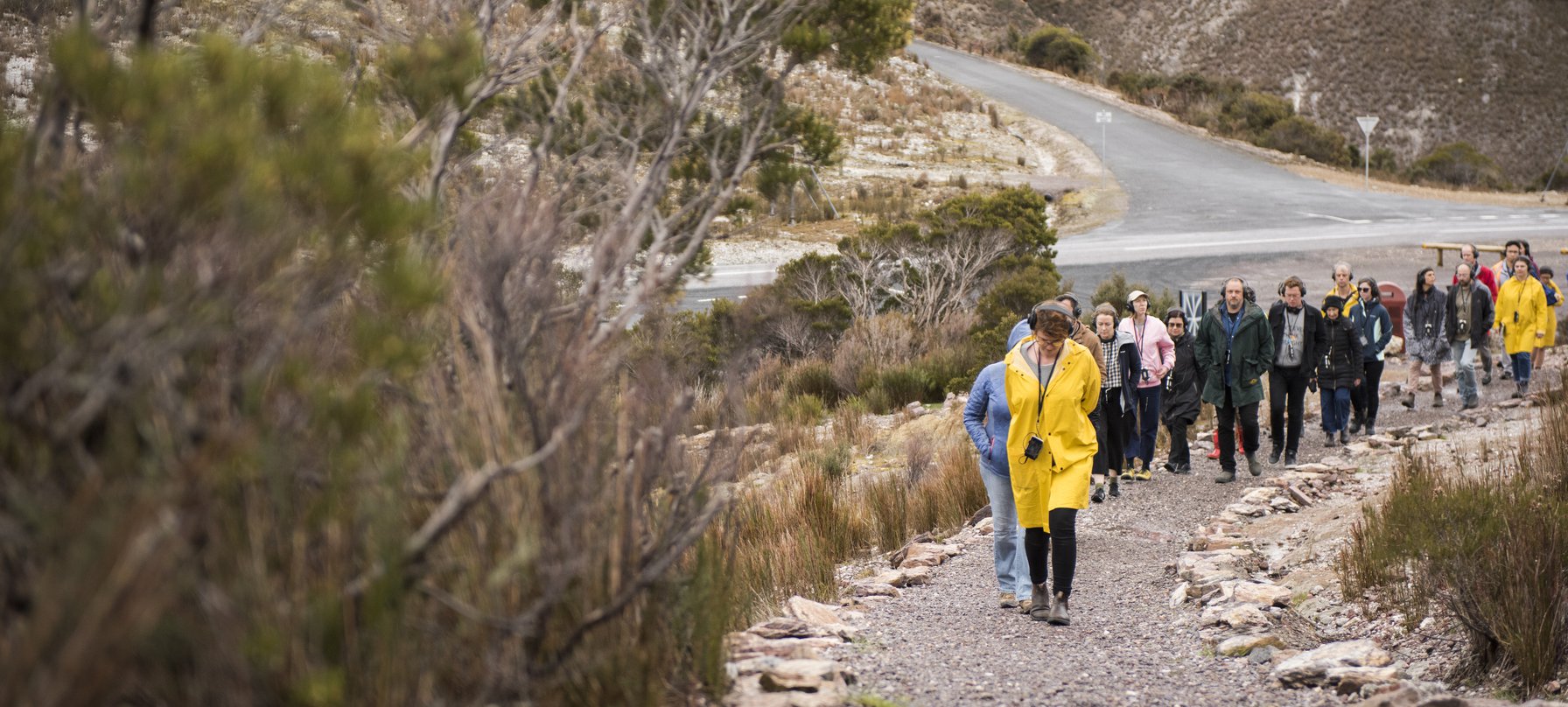 A photo of people walking along a gravel path towards the camera, following a person wearing a vibrant yellow coat. Image by Jack Robert-Tissot.
