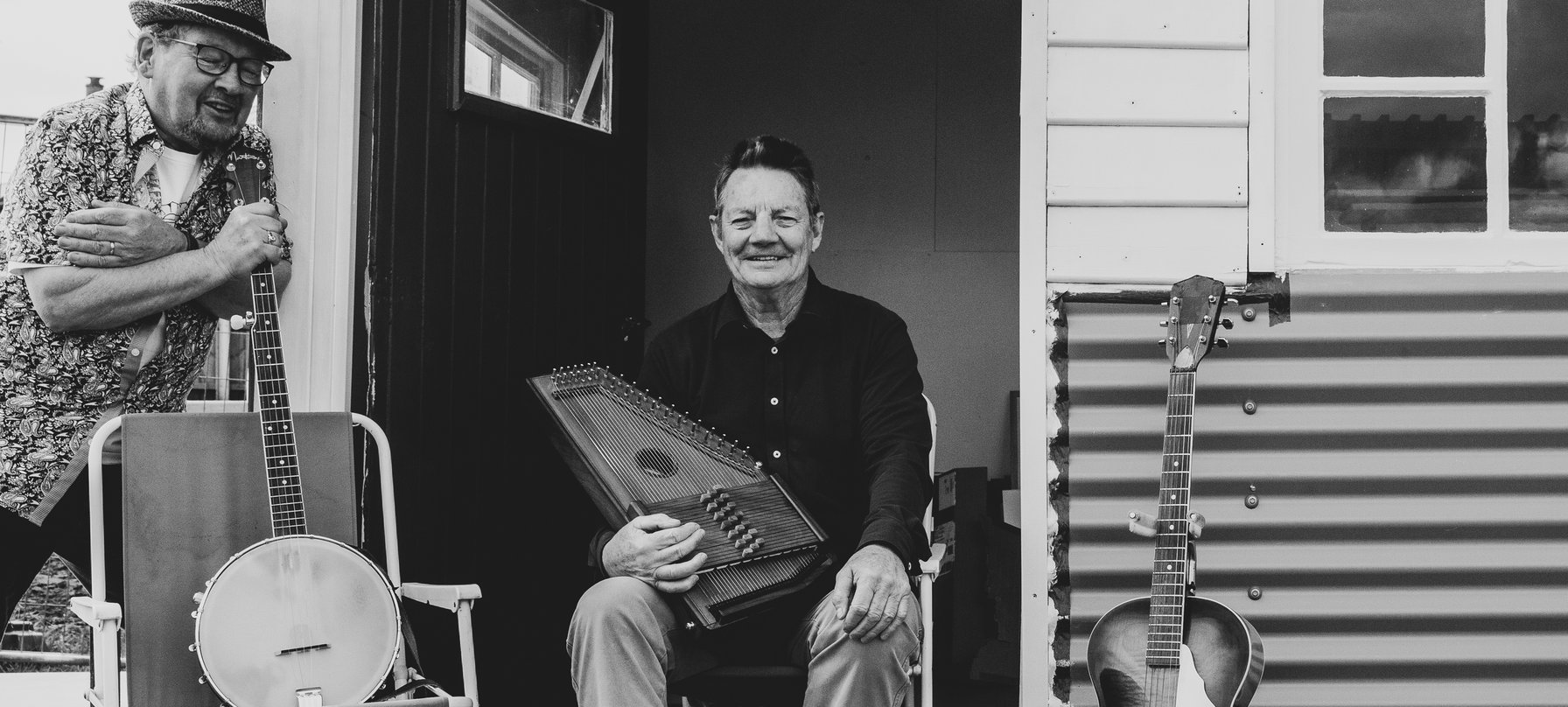 A black and white picture of Tony Newport sitting on a lawn chair on a porch with an autoharp resting on his lap. Tony is surrounded by other instruments and is smiling broadly at the camera. Credit courtesy of artist.