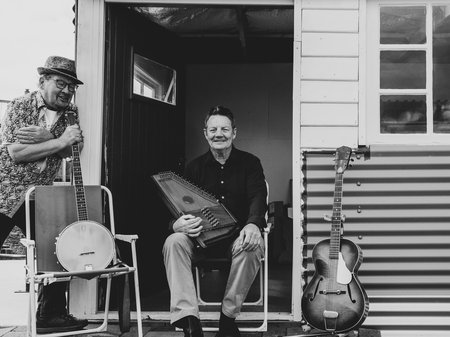 A black and white picture of Tony Newport sitting on a lawn chair on a porch with an autoharp resting on his lap. Tony is surrounded by other instruments and is smiling broadly at the camera. Credit courtesy of artist.