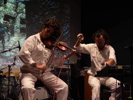Artists Bing / Santospirito x Woods wear white lab coats as they perform, with one playing the violin and the other mixing. An abstract video projects in the background of the performance space.