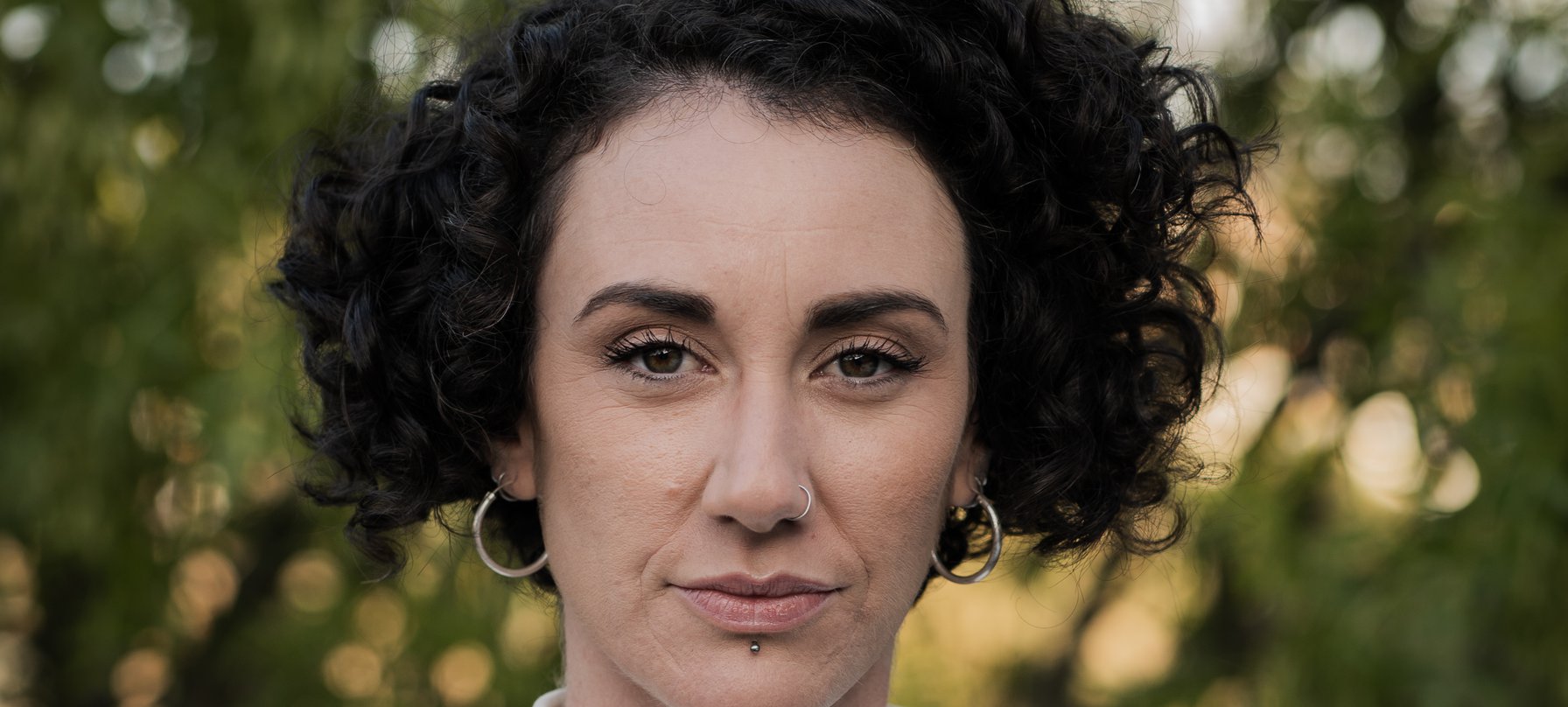 A photo of Frankie Snowdon staring confidently at the camera. Frankie has strong eyebrows, pale skin, a tight curly dark bob haircut and is wearing thick gold hoop earrings. Credit Gabriel Comerford.