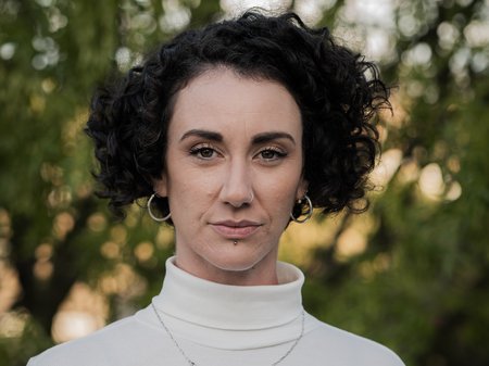 A photo of Frankie Snowdon staring confidently at the camera. Frankie has strong eyebrows, pale skin, a tight curly dark bob haircut and is wearing thick gold hoop earrings. Credit Gabriel Comerford.