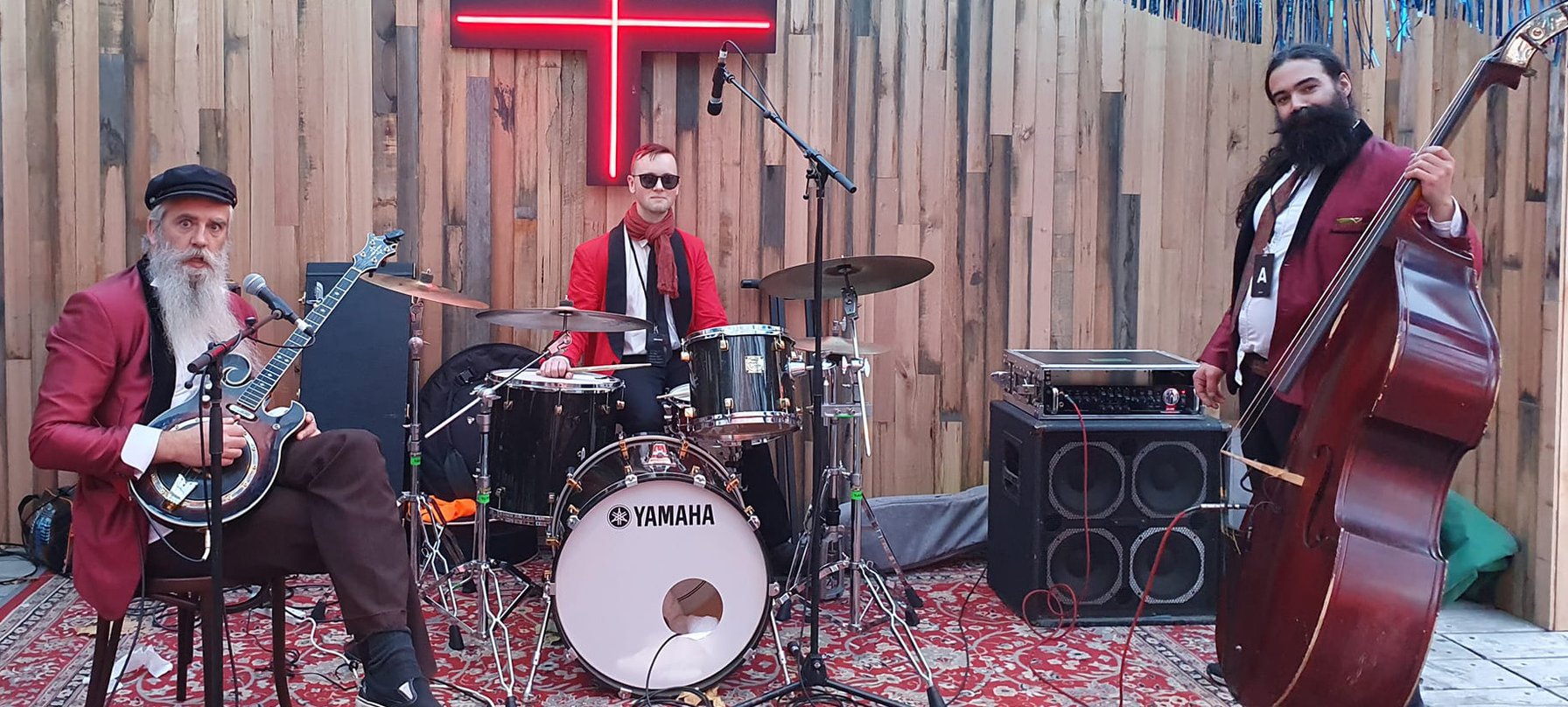 A photo of three people surrounded by musical instruments. A drum kit, electric banjo, double bass and amps can be seen with the musicians who stand on a Persian rug and are all wearing red jackets. Credit