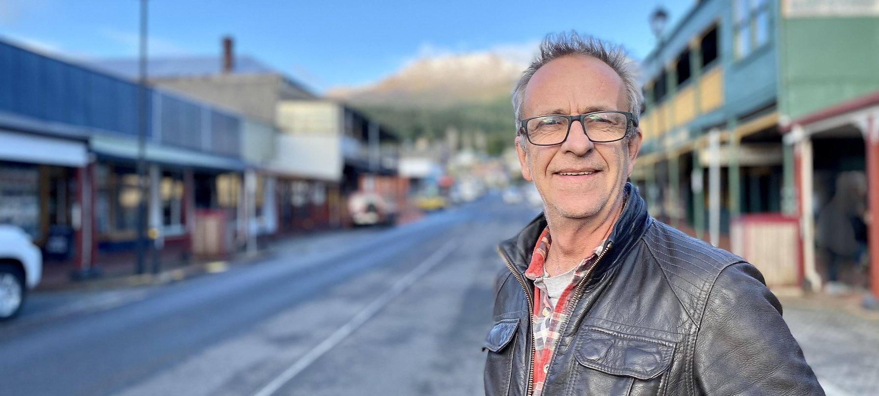 A photo of David "Fitzy" Fitzpatrick standing on Orr Street, Queenstown. Fitzy is smiling and wearing a flannelette shift under a leather jacket.
