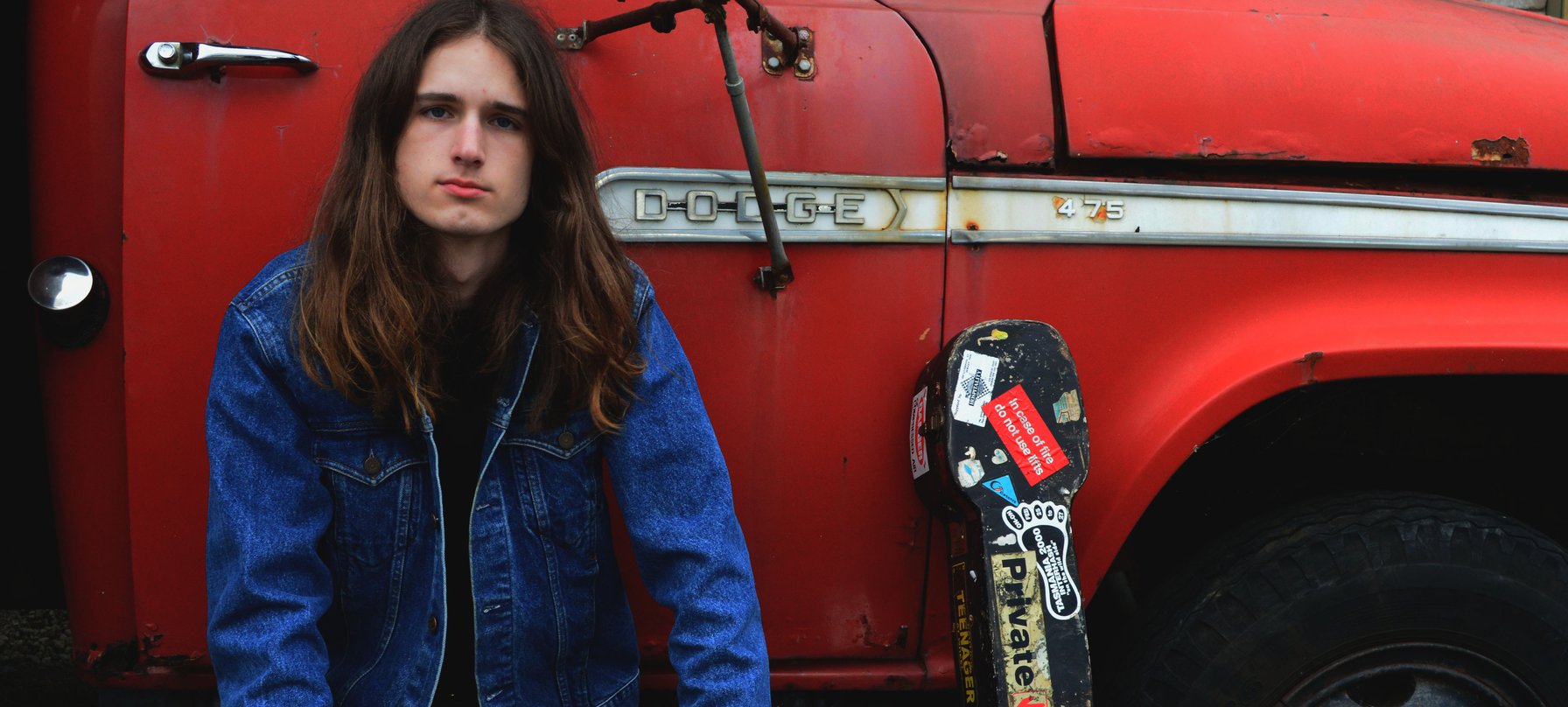 A photo of Eli Strutt sitting on the step of an old red ute. Eli has pale skin and long brown hair past his shoulder. Eli is wearing blue jeans and a blue denim jacket.