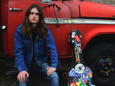 A photo of Eli Strutt sitting on the step of an old red ute. Eli has pale skin and long brown hair past his shoulder. Eli is wearing blue jeans and a blue denim jacket.