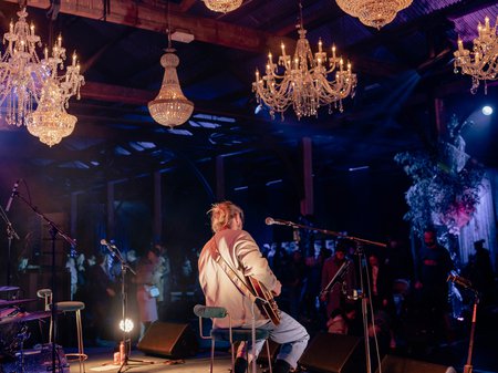 A guitarist of Je Bahl sits atop a stool and plays to an audience. Above the stage are 7 grand chandeliers that glow in the darkness of the night. Credit Mell Schmeider.