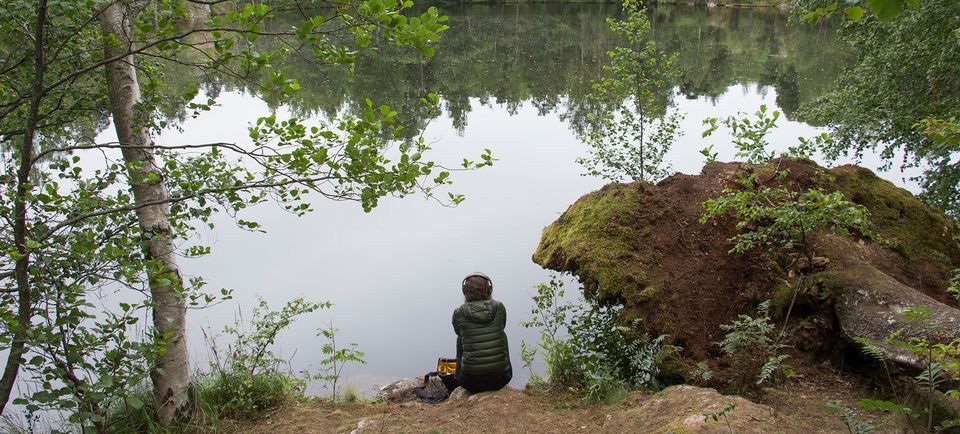 A photo of Polly Stanton sitting on the bank of a lake with their knees pulled up tot heir chest. Lush green trees line the edge of the lake and reflect on the still water.