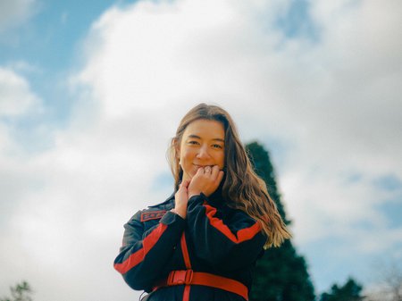 A photo of エミエミ (emi emi) against a cloudy blue sky. emi emi looks down at the camera with her fist up to her chin. She has wavy brown hair past her shoulders, pale skin and is wearing a navy boilersuit with bright red piping and belt. Credit Thomas Wood