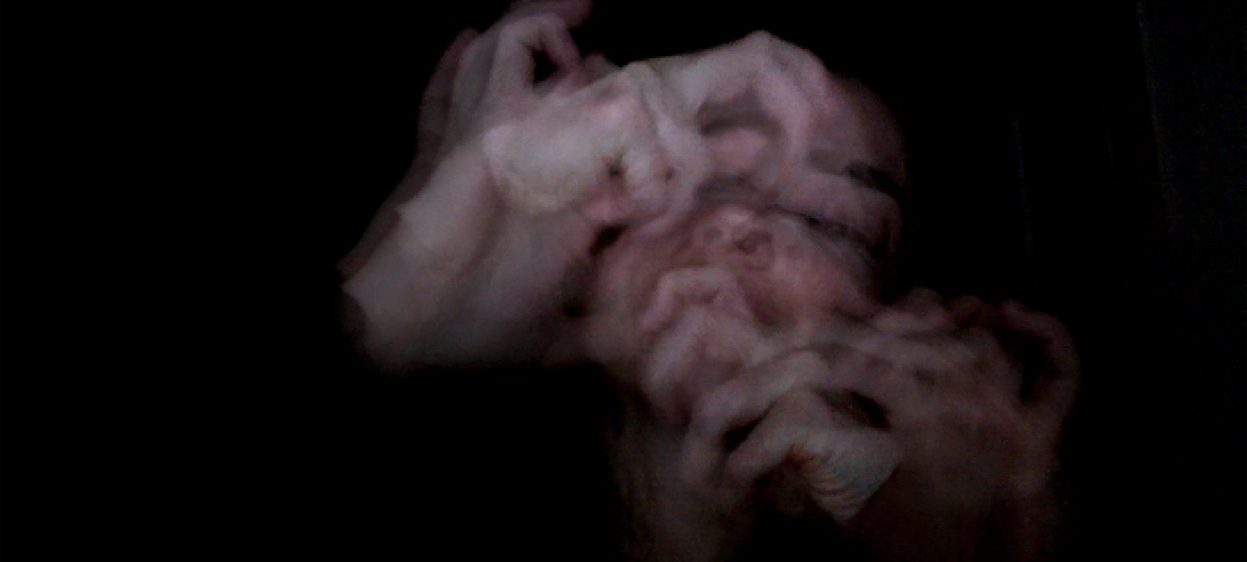 A film still of Sara Wright's face and fingers, blurred, and repeatedly layered over top of themselves, emerging from a black background. Credit Sara Wright.