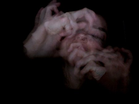 A film still of Sara Wright's face and fingers, blurred, and repeatedly layered over top of themselves, emerging from a black background. Credit Sara Wright.