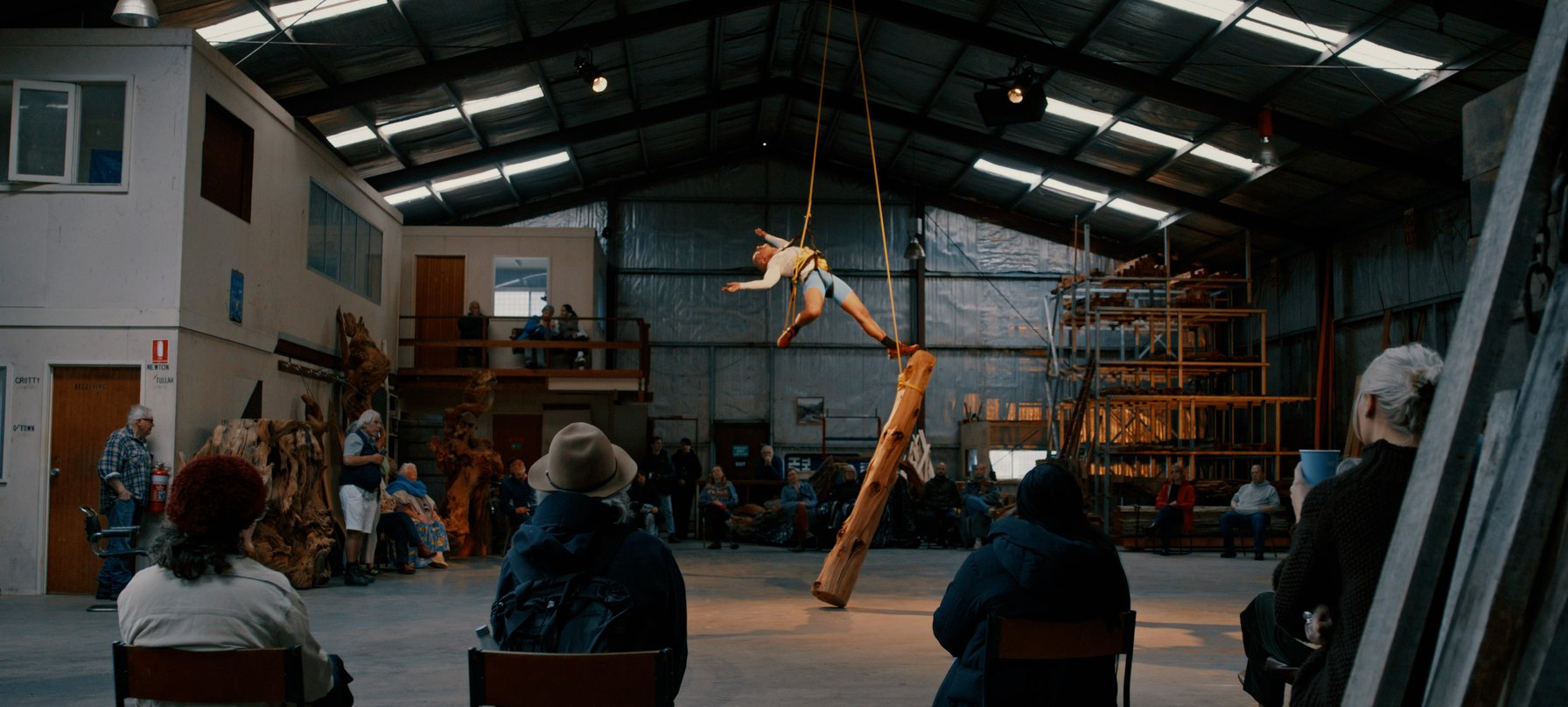 Luke George suspended from the ceiling of a sawmill on a rope with his foot stretched out touching a large wooden log that is vertically suspended beneath him. Still frame from a video documenting the artwork Fell which was presented at The Unconformity.
