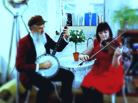 A photo of The Wolfe & Thorn sitting at a small round table with their banjo and violin. Credit courtesy of the artists.